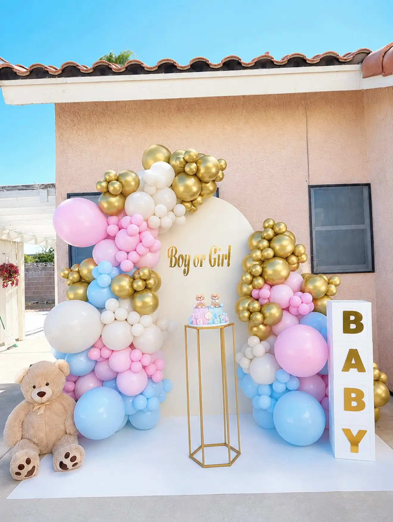 Pastel pink, blue and gold balloon arch with a Boy or Girl sign, teddy bear and BABY blocks for a gender reveal