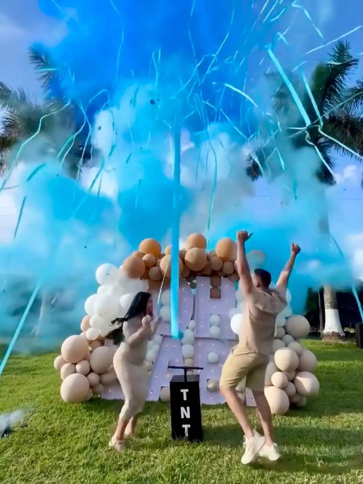 Couple jumping with excitement as a blue smoke cannon reveals a baby boy at an outdoor gender reveal