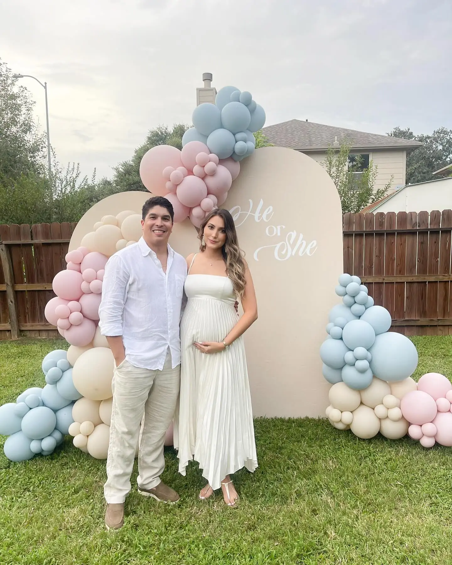 Expecting couple in front of a neutral He or She gender reveal arch with beige, blush and blue balloons
