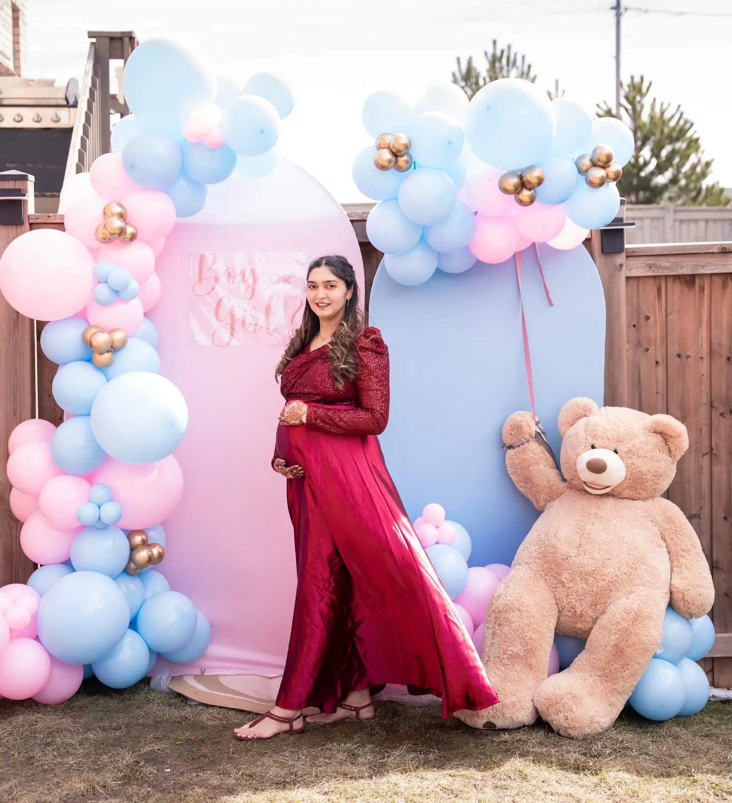 Mother-to-be posing in front of a pink and blue double arch gender reveal backdrop with a giant teddy bear